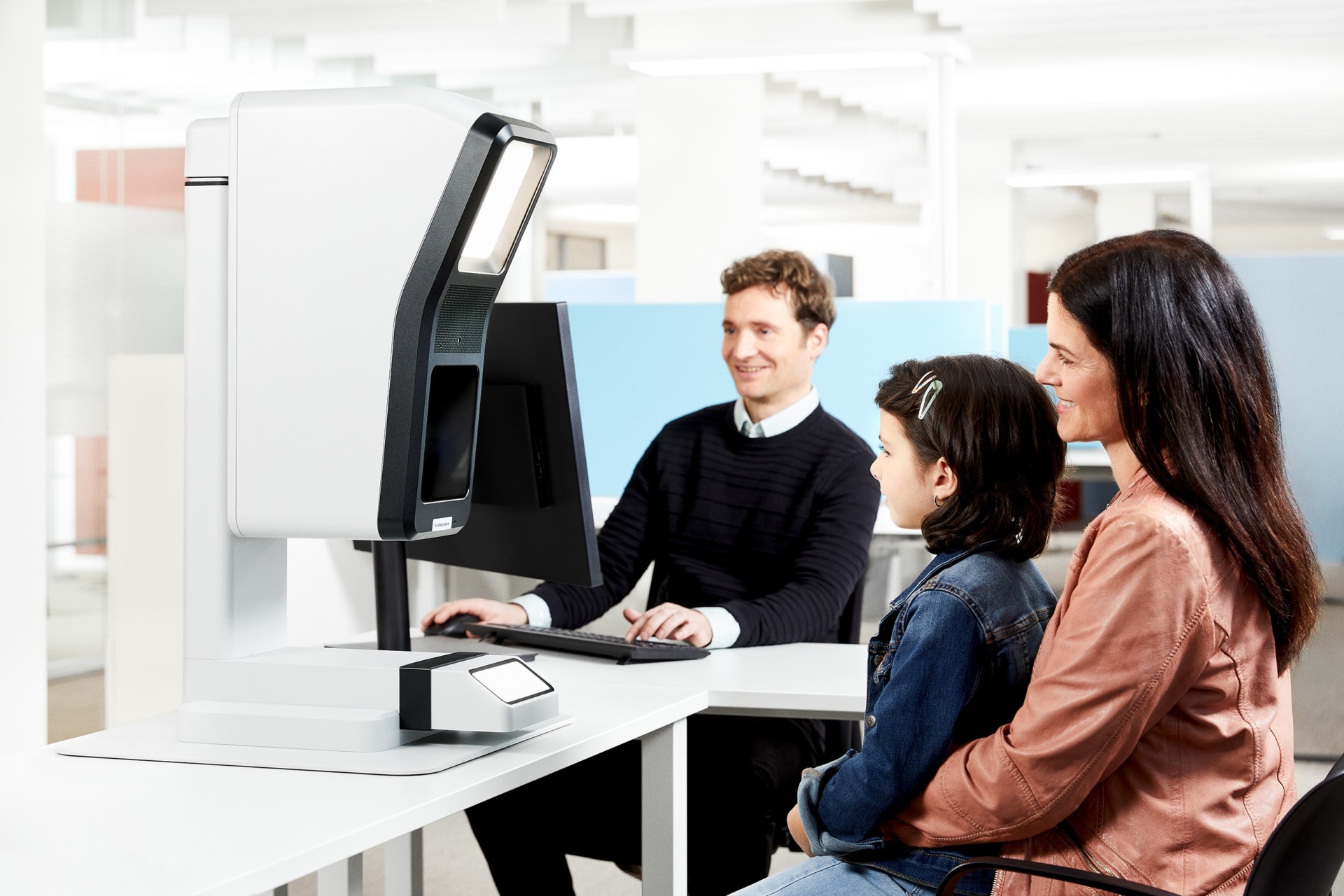 Frau und Kind sitzen vor einem Aufnahmegerät, im Hintergrund ein Mann am PC