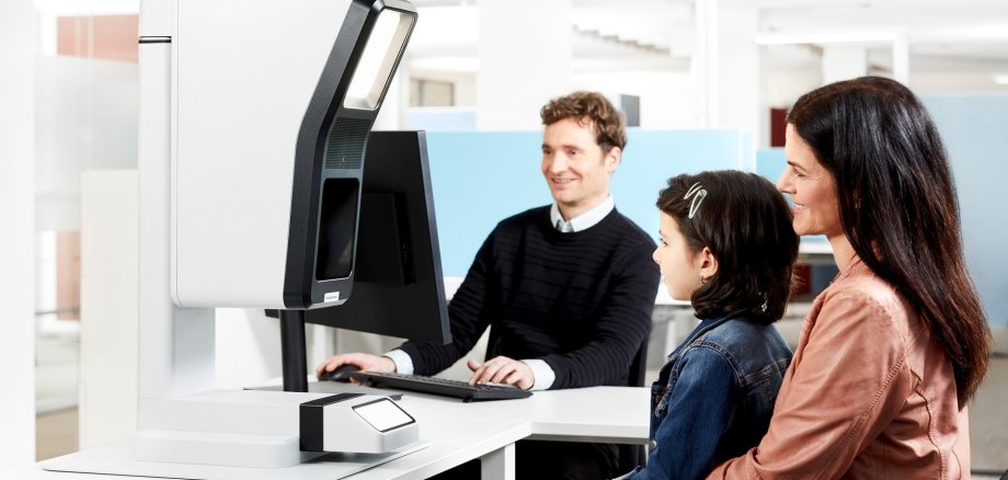 Frau und Kind sitzen vor einem Aufnahmegerät, im Hintergrund ein Mann am PC
