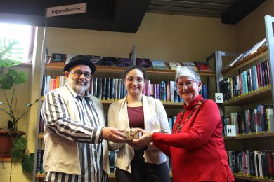 Spendenübergabe Jugendlounge Drei Personen halten eine Spendenbox, im Hintergrund Bücher