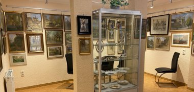 Kulturstube - Franz Huth Bildergalerie Vitrine mit Porzellantellern und -krügen, dahinter Wand mit Gemälden