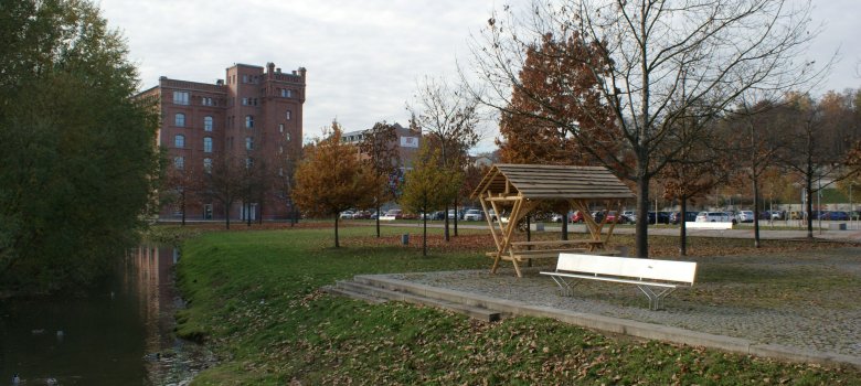 befestigter Platz neben einem kleinen Fluss, im Hintergrund Backsteingebäude und Parkplatz